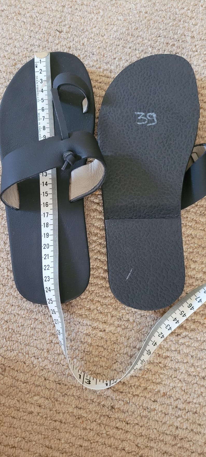 May include: A pair of black sandals with a toe loop and a knotted strap design. One sandal shows the number 39 written on the footbed. A white measuring tape is placed over the sandals, indicating a length of approximately 25 centimeters.
