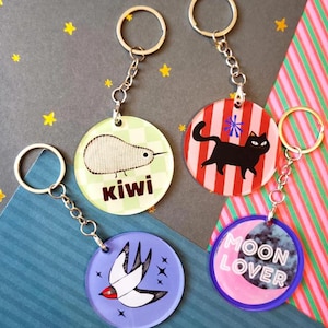 May include: Four round keychains with various designs. One features a kiwi bird with the word "kiwi", another a black cat on red and white stripes, a third a white bird, and the last one says "MOON LOVER". Each keychain has a silver chain and ring.