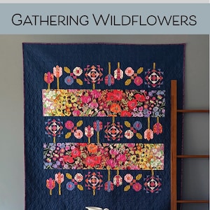 May include: A navy blue quilt with floral patterns and the text "Gathering Wildflowers." The quilt features rows of colourful flowers and geometric shapes. The quilt is displayed on a wall above a wooden table.