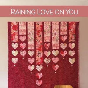 Pode incluir: Um edredom vermelho com um design de coração, pendurado em uma parede. O edredom apresenta painéis florais verticais e fileiras de formas de coração. O texto "RAINING LOVE ON YOU" está no topo. Uma cadeira de madeira está abaixo.