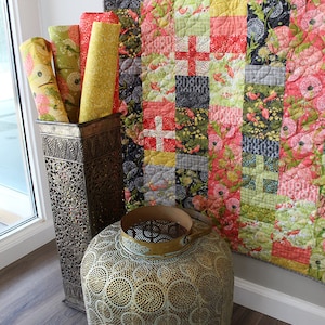 May include: A colorful patchwork quilt with a floral and bird design hangs on a white wall. The quilt is made of various fabrics in shades of pink, red, green, yellow, and black. The quilt is displayed in a room with a wooden floor and a metal decorative stand.