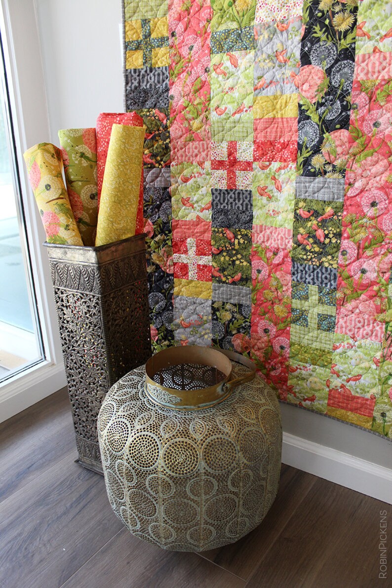 May include: A colorful patchwork quilt with a floral and bird design hangs on a white wall. The quilt is made of various fabrics in shades of pink, red, green, yellow, and black. The quilt is displayed in a room with a wooden floor and a metal decorative stand.