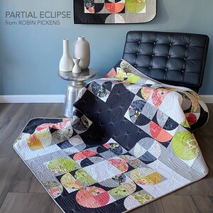 Peut inclure: Une courtepointe moderne avec un motif g&eacute;om&eacute;trique pr&eacute;sentant des motifs en demi-cercle en noir, blanc, gris et diverses couleurs. La courtepointe est drap&eacute;e sur une chaise en cuir noir et sur le sol. Le texte "PARTIAL ECLIPSE from ROBIN PICKENS" est visible.