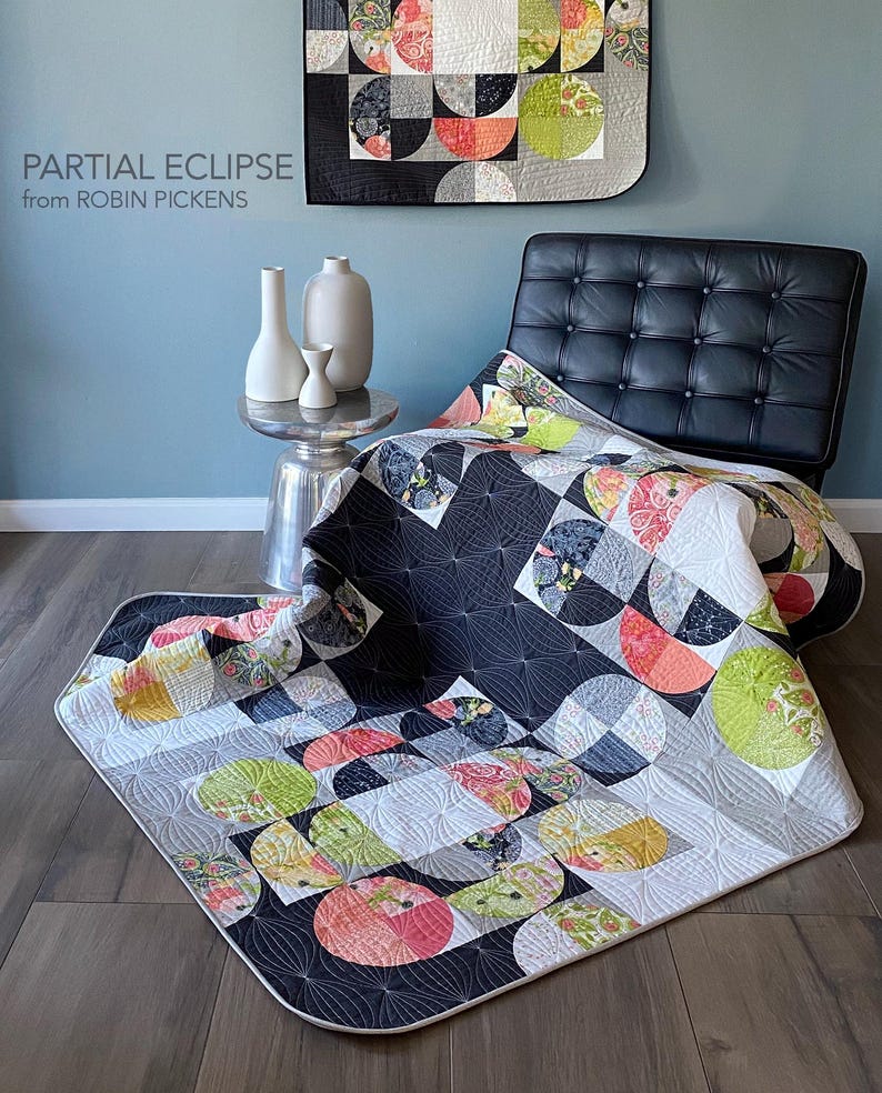 Peut inclure: Une courtepointe moderne avec un motif g&eacute;om&eacute;trique pr&eacute;sentant des motifs en demi-cercle en noir, blanc, gris et diverses couleurs. La courtepointe est drap&eacute;e sur une chaise en cuir noir et sur le sol. Le texte "PARTIAL ECLIPSE from ROBIN PICKENS" est visible.
