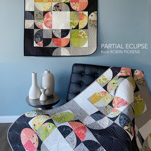 Peut inclure: Une courtepointe color&eacute;e avec un motif g&eacute;om&eacute;trique de demi-cercles aux motifs et couleurs vari&eacute;s, notamment rose, vert et jaune, sur un fond noir et gris. La courtepointe est expos&eacute;e sur un mur et drap&eacute;e sur une chaise. Le texte "PARTIAL ECLIPSE from ROBIN PICKENS" est visible.