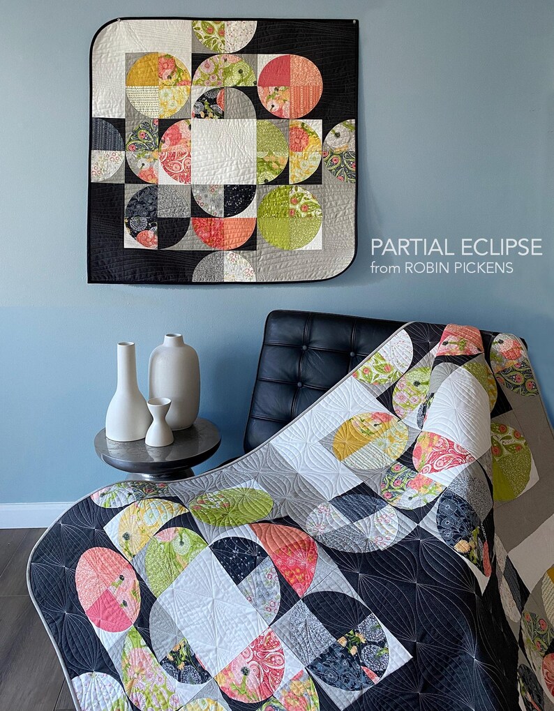 Peut inclure: Une courtepointe color&eacute;e avec un motif g&eacute;om&eacute;trique de demi-cercles aux motifs et couleurs vari&eacute;s, notamment rose, vert et jaune, sur un fond noir et gris. La courtepointe est expos&eacute;e sur un mur et drap&eacute;e sur une chaise. Le texte "PARTIAL ECLIPSE from ROBIN PICKENS" est visible.