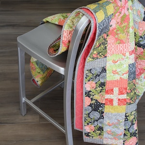 May include: A patchwork quilt with a floral and bird design in shades of pink, green, yellow, and gray. The quilt is folded and draped over a silver metal chair.