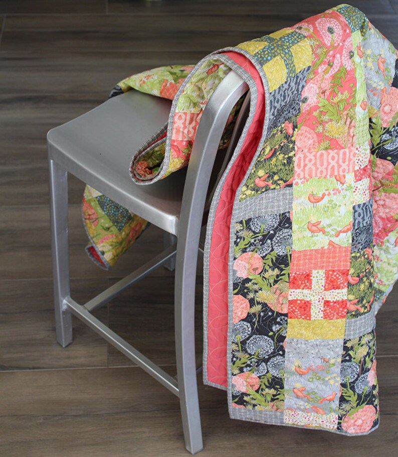 May include: A patchwork quilt with a floral and bird design in shades of pink, green, yellow, and gray. The quilt is folded and draped over a silver metal chair.