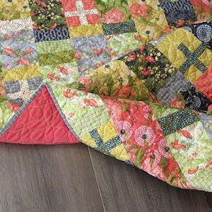 May include: A patchwork quilt with a floral and bird print. The quilt is made of various fabrics in shades of green, red, yellow, and gray. The quilt is folded and lying on a wooden floor.