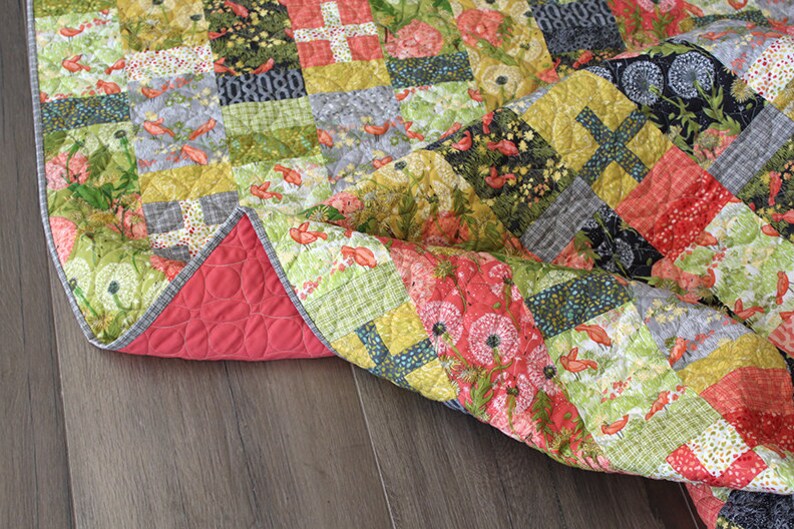 May include: A patchwork quilt with a floral and bird print. The quilt is made of various fabrics in shades of green, red, yellow, and gray. The quilt is folded and lying on a wooden floor.