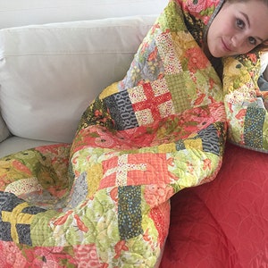 May include: A colorful patchwork quilt with a variety of floral and geometric patterns in shades of pink, green, yellow, and gray. The quilt is draped over a white couch and a red throw pillow.