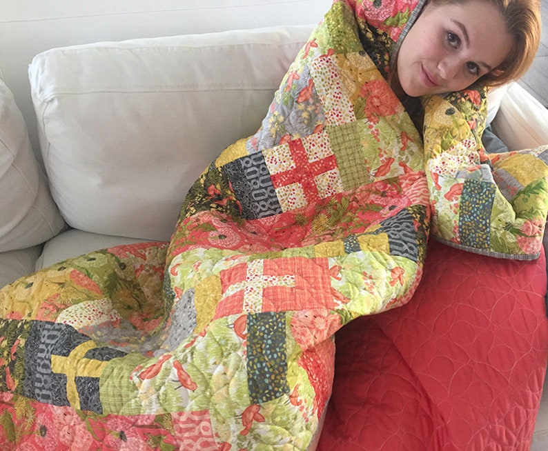 May include: A colorful patchwork quilt with a variety of floral and geometric patterns in shades of pink, green, yellow, and gray. The quilt is draped over a white couch and a red throw pillow.