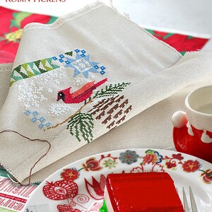 May include: A white fabric with a cross-stitch design of a cardinal bird on a pine branch with snow. The fabric is folded and placed on a table with a red and white patterned plate and a red and white dessert. The text "SEASONAL COURIER: CARDINAL'S WINTER ROBIN PICKENS" is at the top of the image.