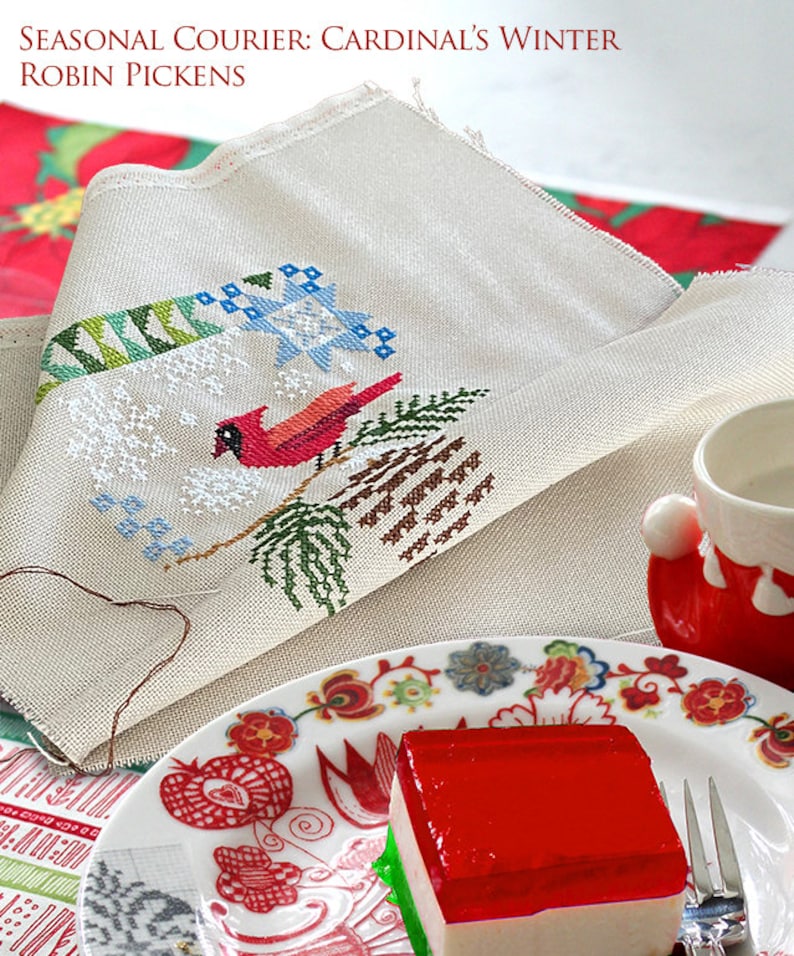 May include: A white fabric with a cross-stitch design of a cardinal bird on a pine branch with snow. The fabric is folded and placed on a table with a red and white patterned plate and a red and white dessert. The text "SEASONAL COURIER: CARDINAL'S WINTER ROBIN PICKENS" is at the top of the image.