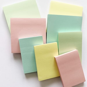 May include: A collection of pastel-coloured sticky note pads in shades of pink, green, yellow, and blue. The pads are rectangular and arranged in a layered composition, with some overlapping others. The image is shot from above, against a white background.