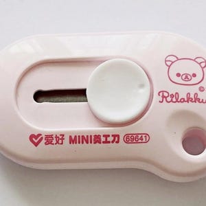 May include: A pale pink utility knife with a white circular slider and a small blade. The knife features a cartoon bear illustration and the text "Rilakkuma" and "San-X". Additional text includes Chinese characters and the number "69641".