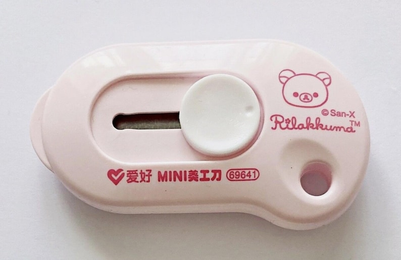 May include: A pale pink utility knife with a white circular slider and a small blade. The knife features a cartoon bear illustration and the text "Rilakkuma" and "San-X". Additional text includes Chinese characters and the number "69641".