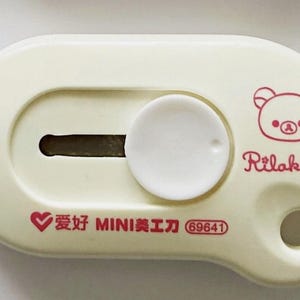 May include: A cream-coloured utility knife with a white circular slider and a blade slot. The design includes a red illustration of a bear and the text "Rilakkuma" and "San-X". Additional text in red includes Chinese characters and the number "69641".