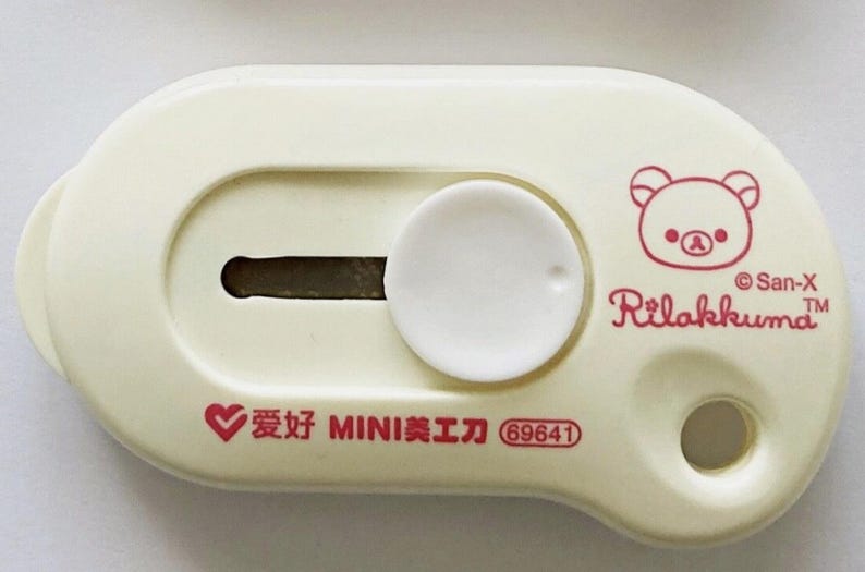 May include: A cream-coloured utility knife with a white circular slider and a blade slot. The design includes a red illustration of a bear and the text "Rilakkuma" and "San-X". Additional text in red includes Chinese characters and the number "69641".