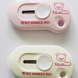 May include: Two small, pastel-coloured utility knives. One is light pink, the other is cream. Each has a sliding blade, a white circular button, and a Rilakkuma bear logo. The text "爱好 MINI美工刀 69641" is printed on each.