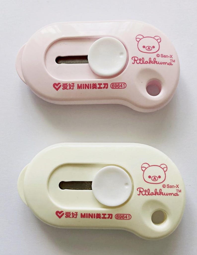 May include: Two small, pastel-coloured utility knives. One is light pink, the other is cream. Each has a sliding blade, a white circular button, and a Rilakkuma bear logo. The text "爱好 MINI美工刀 69641" is printed on each.