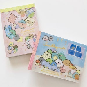 May include: Two pink and blue memo pads featuring Sumikko Gurashi characters. The memo pads have a pastel colour scheme and feature a variety of cute characters. The text on the memo pads reads "Sumikkogurashi".