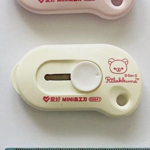 May include: Two Rilakkuma-themed craft knives in pink and cream, each with a circular blade cover and a white button. The pink knife has the text "爱好 MINI美工刀 69641" and a bear logo. A ruler with cartoon characters is at the bottom.