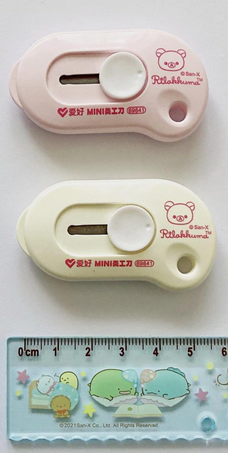 May include: Two Rilakkuma-themed craft knives in pink and cream, each with a circular blade cover and a white button. The pink knife has the text "爱好 MINI美工刀 69641" and a bear logo. A ruler with cartoon characters is at the bottom.