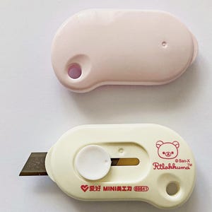 May include: Two small utility knives. One is pale pink, the other is off-white with a brown blade. The off-white knife has a Rilakkuma bear graphic and text. The pink knife has a hole on one end.