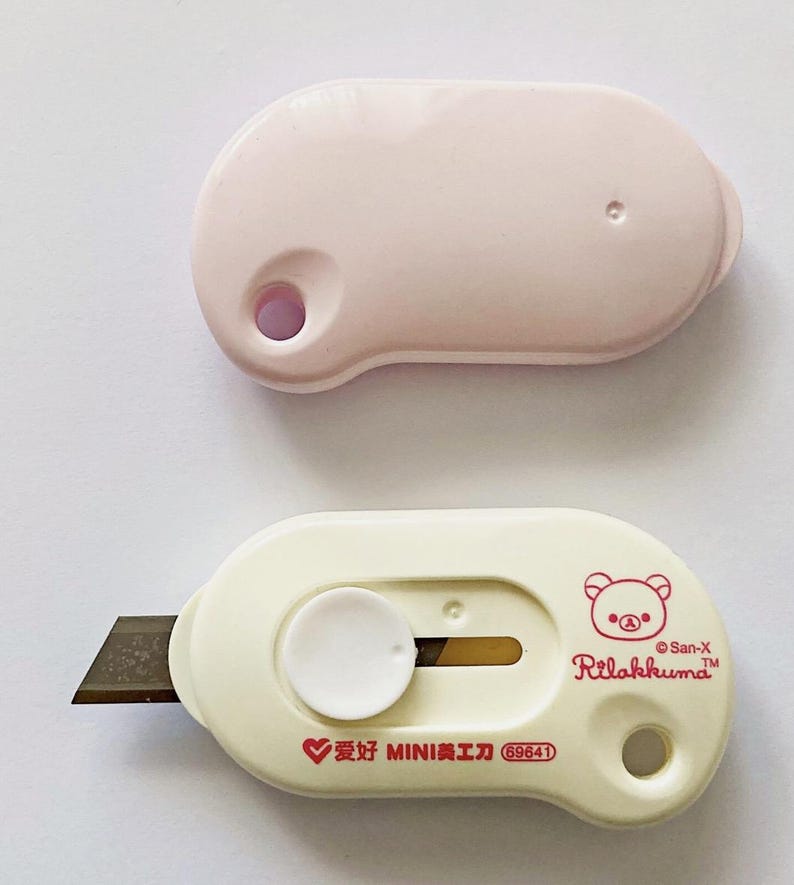 May include: Two small utility knives. One is pale pink, the other is off-white with a brown blade. The off-white knife has a Rilakkuma bear graphic and text. The pink knife has a hole on one end.