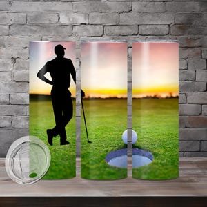 Puede incluir: Un juego de tres vasos de acero inoxidable con una silueta de golfista y una bola de golf en un hoyo en un campo de golf verde. El fondo es un cielo de puesta de sol.