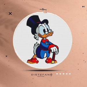 Puede incluir: Un aro de bordado blanco con una imagen de dibujos animados de un pato con un sombrero de copa y un bastón. El pato lleva una chaqueta azul con detalles rojos. El fondo es blanco.