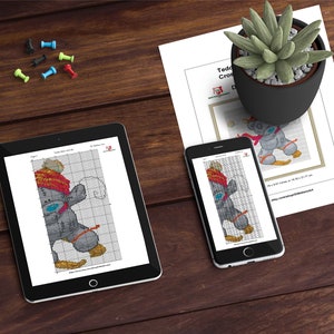 Peut inclure: Un motif de point de croix num&eacute;rique pour un ours en peluche gris portant un chapeau rouge et blanc et tenant une boule de neige. Le motif est affich&eacute; sur une tablette et un smartphone.