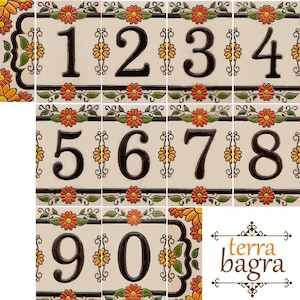 Pode incluir: Um conjunto de dez números de casa de cerâmica com um design floral. Os números são pretos e o fundo é branco com flores laranja e amarelas. Os números estão dispostos em uma grade. O canto inferior direito da imagem mostra o texto "terra bagra".