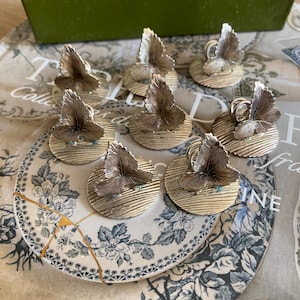 May include: Eight gold-coloured leaf-shaped napkin rings. Each ring has a detailed leaf design on a circular base. The rings are displayed on a floral patterned plate, suggesting a decorative table setting.