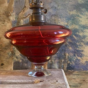 Peut inclure: Une lampe à huile en verre rouge avec un brûleur en métal et une base ronde à plusieurs niveaux. La lampe est posée sur une surface en bois.