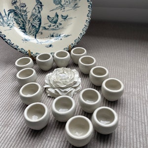 Peut inclure: Une collection de 13 tasses en céramique blanche disposées en cercle autour d'une fleur en céramique blanche. Les tasses sont toutes de la même taille et de la même forme, et elles ont une finition lisse et brillante.