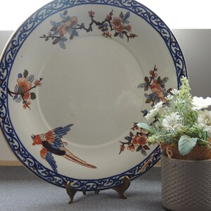 May include: A large white ceramic plate with a blue and gold rim and a floral design featuring a bird. The plate is on a stand and a small potted plant is in the foreground.