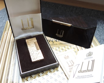 May include: A gold Dunhill lighter in its original box. The lighter has a textured design and is made of metal. The box is black with a gold trim and the Dunhill logo.