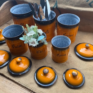 Peut inclure: Ensemble de pots de cuisine orange et noirs avec couvercles. Les pots ont un intérieur en métal galvanisé et sont étiquetés avec des mots français. Certains pots contiennent des ustensiles et des fleurs.