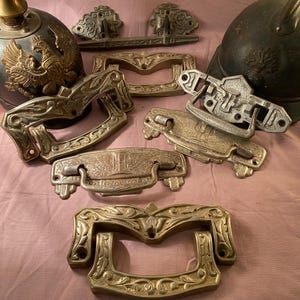 Puede incluir: Una variedad de herrajes metálicos antiguos, que incluyen tiradores de cajones ornamentados y cascos decorativos. Los tiradores de cajones presentan intrincados diseños florales y de volutas en varios tamaños y acabados. Los cascos son oscuros con detalles en relieve.