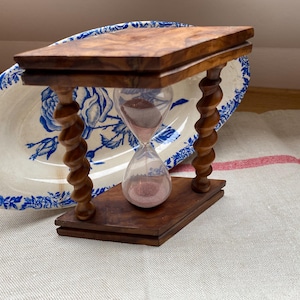 Puede incluir: Un reloj de arena de madera con acabado marrón y una bombilla de vidrio transparente llena de arena rosa. El reloj de arena está sostenido por una base rectangular y tiene columnas retorcidas talladas intrincadamente. Un plato con estampado floral azul y blanco está en el fondo.