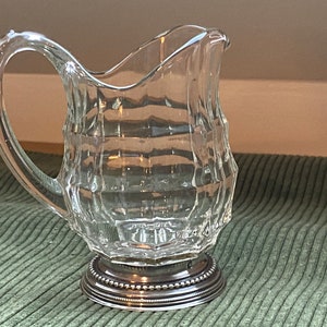 May include: A clear glass pitcher with a silver base. The pitcher has a textured, faceted design and a rounded handle.