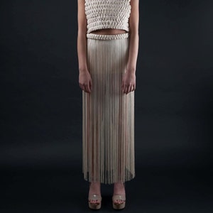 May include: A cream-colored, two-piece macrame outfit. The top is a square-neck crop top with thick straps. The skirt is a long, fringed design. The model is wearing platform heels. The background is a solid black.