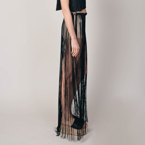 May include: Black fringe crop top and skirt set. The top has thin straps and a fringed hem. The skirt is sheer with long fringe. The outfit is worn with black heels. The background is plain white.