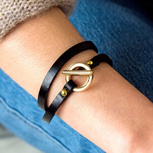 May include: A black leather wrap bracelet with a gold-toned toggle clasp. The bracelet is worn on a person's wrist.