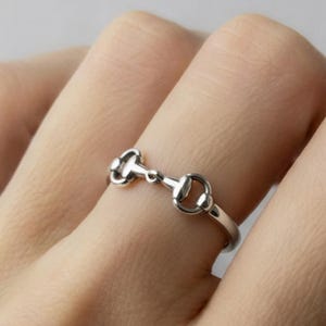 Può includere: Un anello color argento con un design a morso di cavallo. L'anello è indossato su un dito e ha un'estetica delicata ispirata all'equitazione. Il design dell'anello include due estremità arrotondate collegate da una barra centrale.