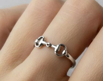 Puede incluir: Un anillo plateado con un diseño de bocado de caballo. El anillo se lleva en un dedo y tiene una estética delicada inspirada en la equitación. El diseño del anillo incluye dos extremos redondeados conectados por una barra central.