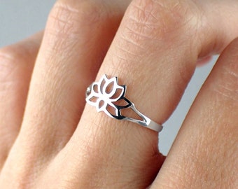 Puede incluir: Un anillo de plata con un delicado diseño de flor de loto. El anillo se lleva en el dedo medio de una mano.