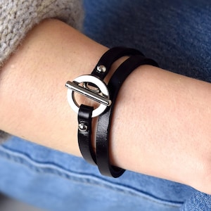May include: A black leather wrap bracelet with a silver circular clasp. The bracelet is worn on a person's wrist.